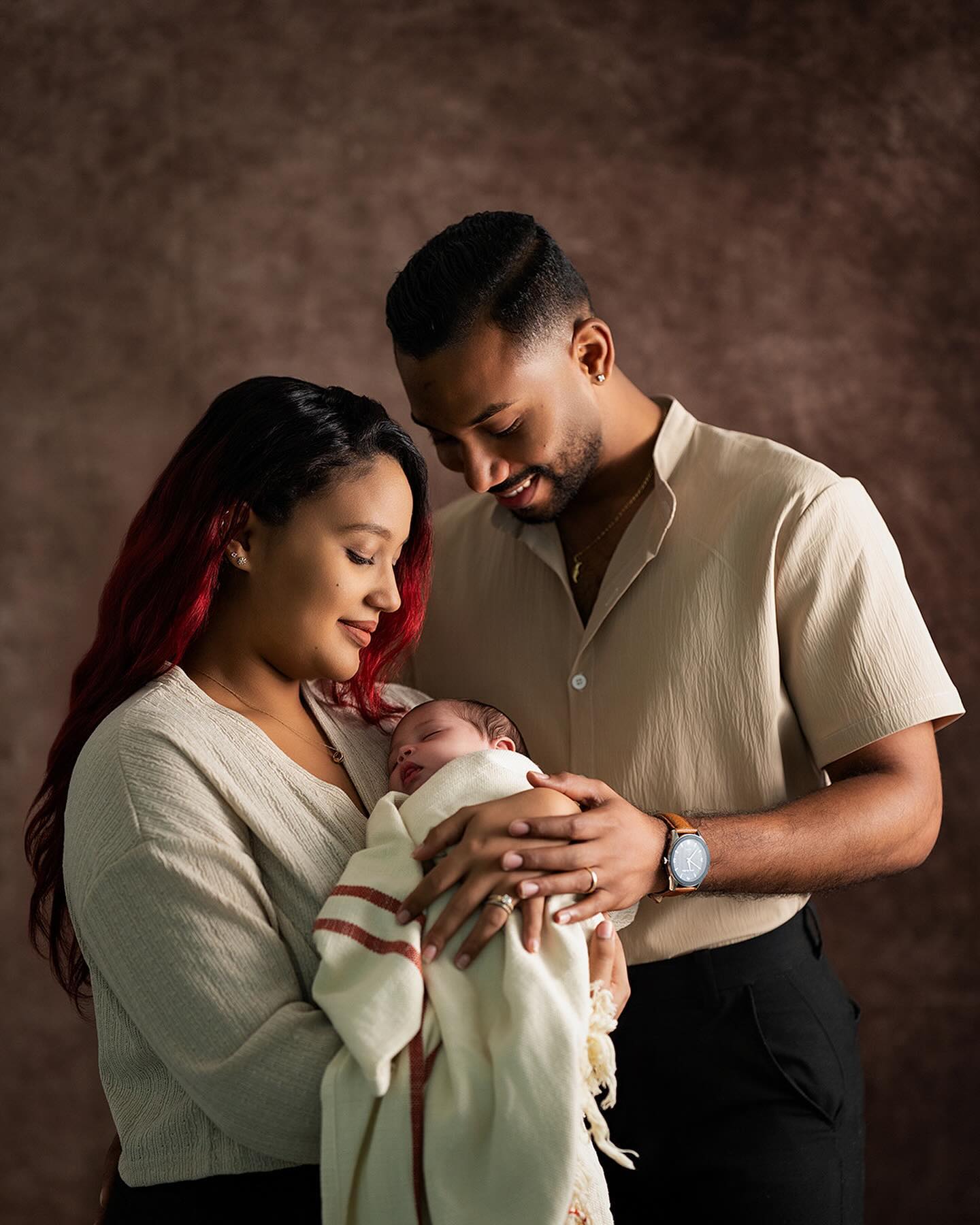 Loving couple cradles their newborn baby in a cozy studio setting symbolizing family warmth and love with parents gazing down fondly at their sleeping child.