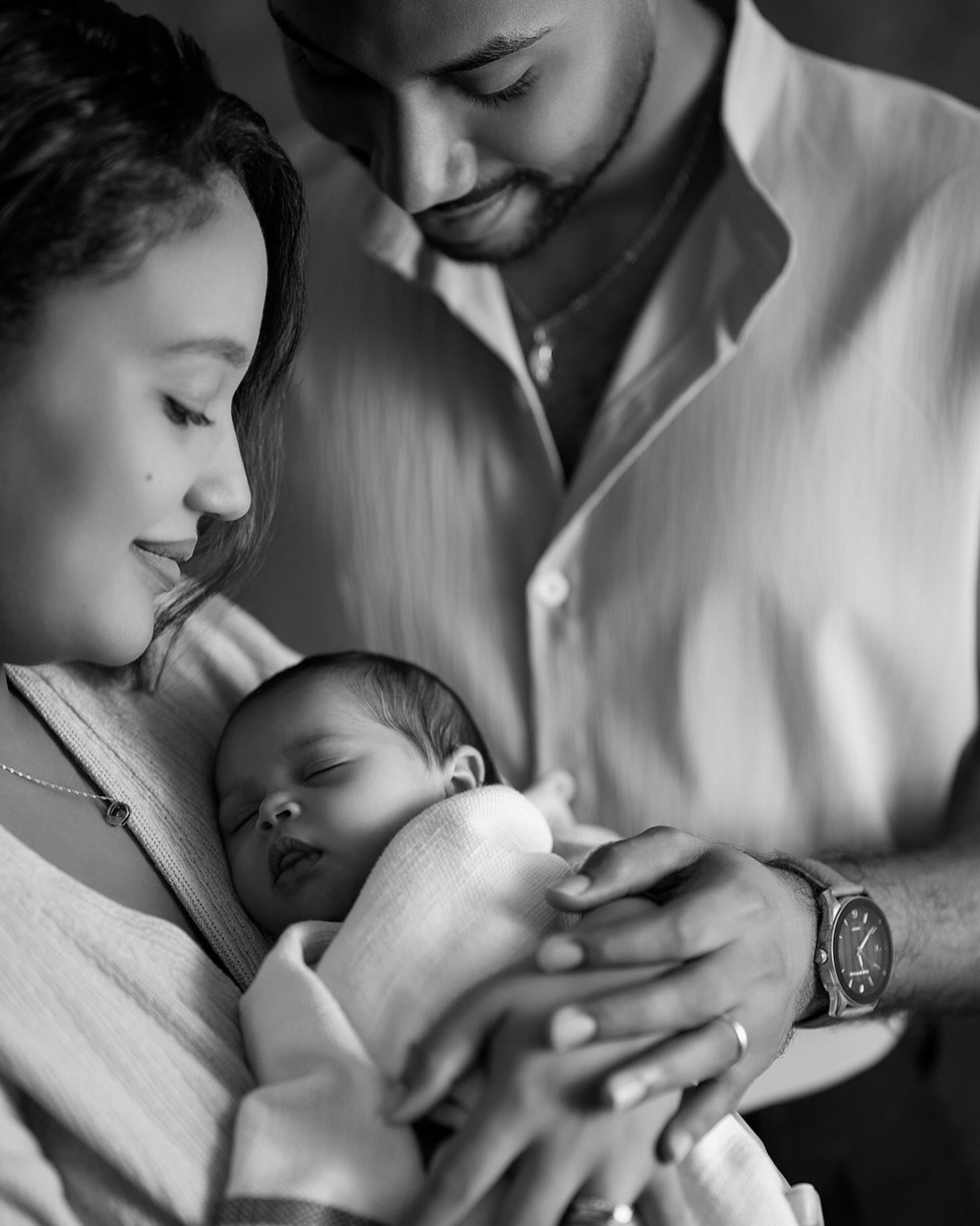 Loving parents gently cradle their sleeping newborn wrapped in a soft blanket. The intimate black and white portrait captures a tender family moment filled with warmth.