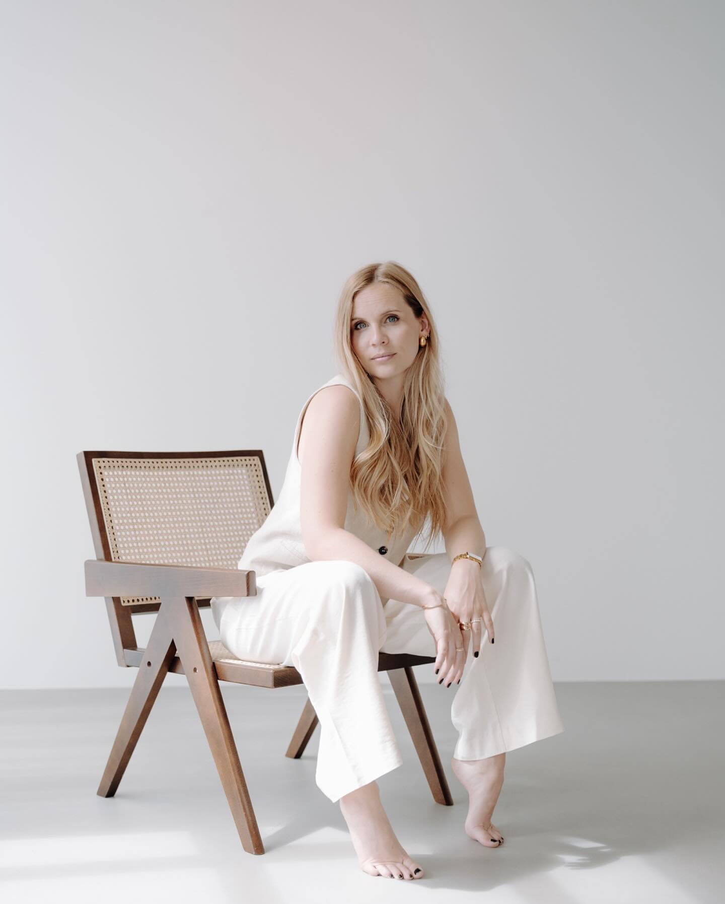 Young woman with long blonde hair sitting on a modern wooden chair wears a stylish white outfit against a minimalist background exuding elegance and calmness.