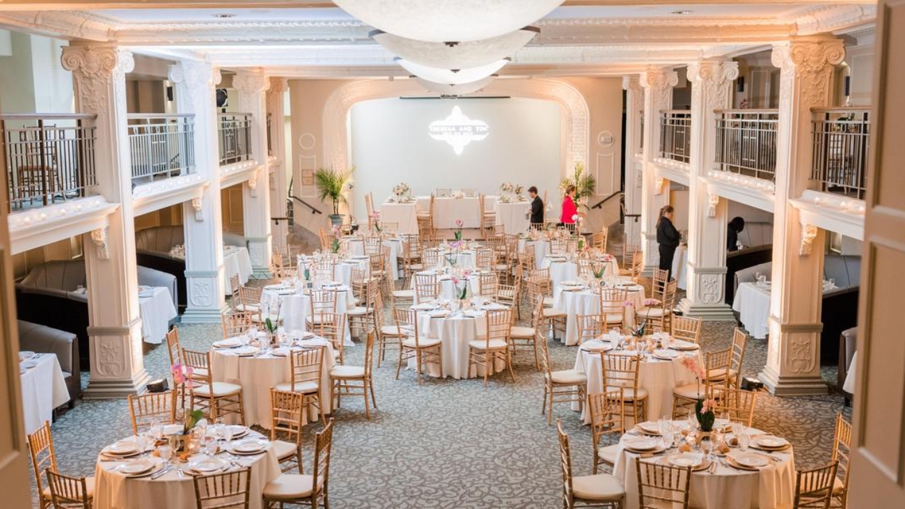 Elegant banquet hall with round tables set for a wedding reception featuring white tablecloths and gold chairs under a grand chandelier for a stylish celebration.