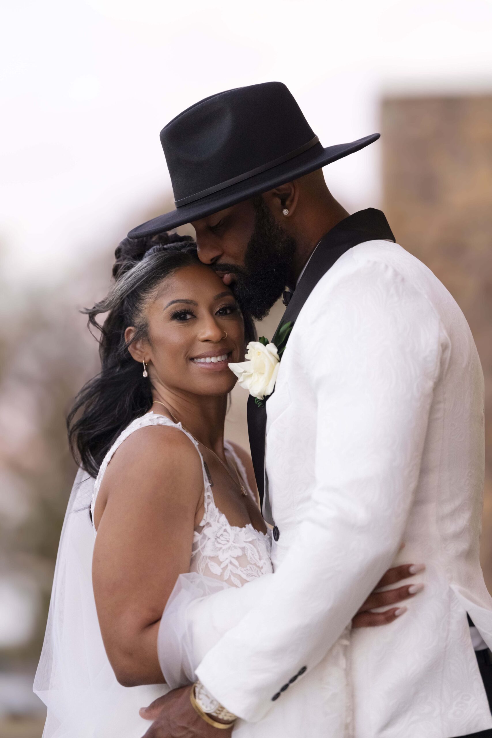 Bride and groom embracing on their wedding day with the groom in a white suit and black hat and the bride in a lace gown smiling against a blurred outdoor background.