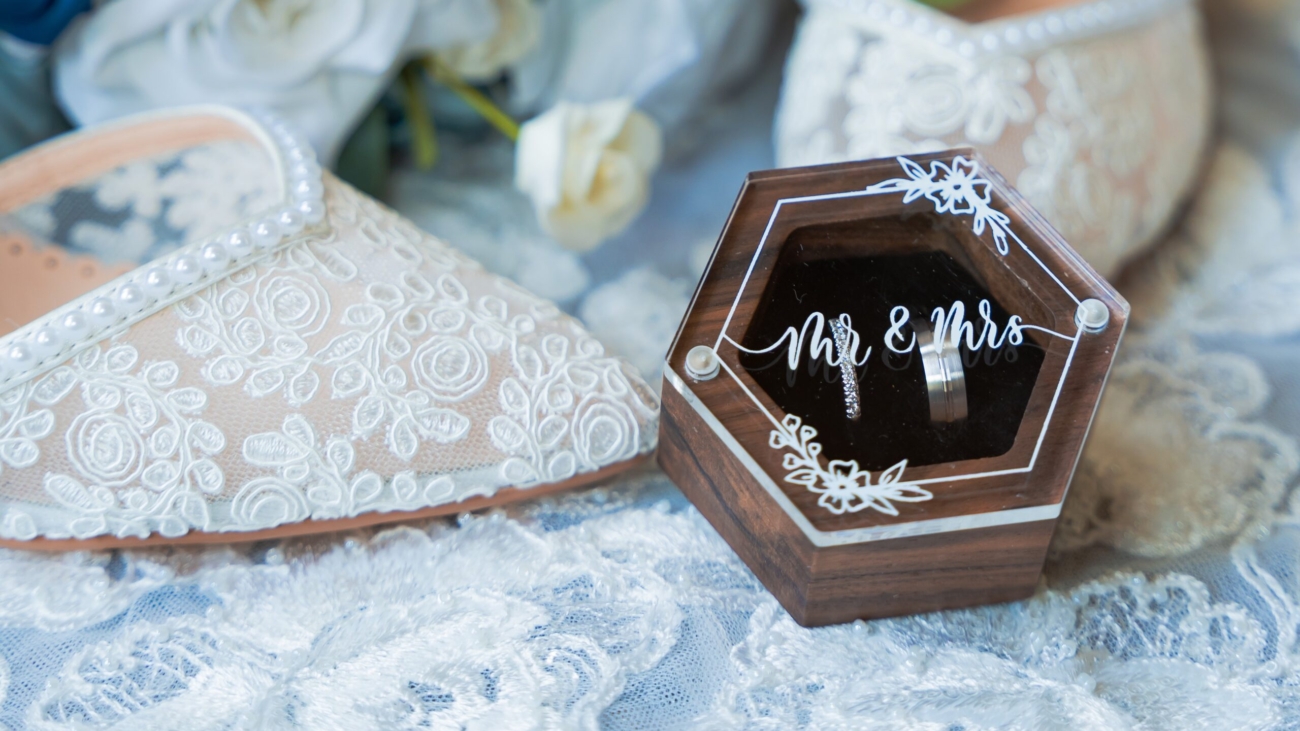 Elegant wedding scene with lace bridal shoes next to a wooden hexagon ring box labeled Mr and Mrs surrounded by white and blue flowers on a delicate lace background