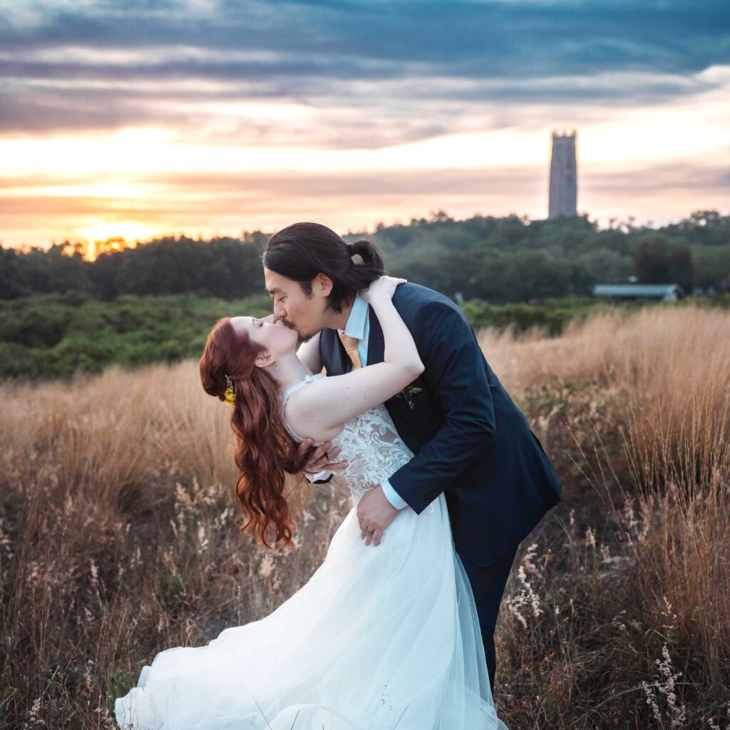 A couple shares a romantic kiss in a scenic outdoor landscape at sunset, surrounded by tall grass with a distant tower in the background, capturing a beautiful moment.