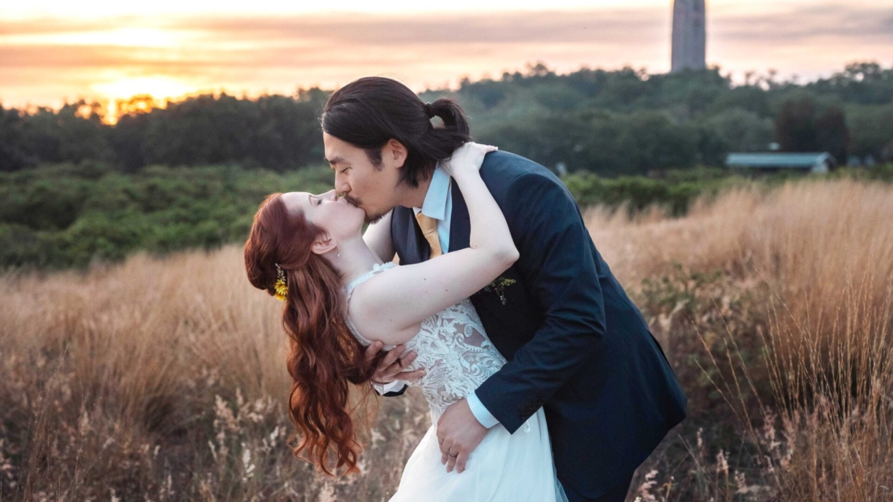 A couple shares a romantic kiss in a scenic outdoor landscape at sunset, surrounded by tall grass with a distant tower in the background, capturing a beautiful moment.