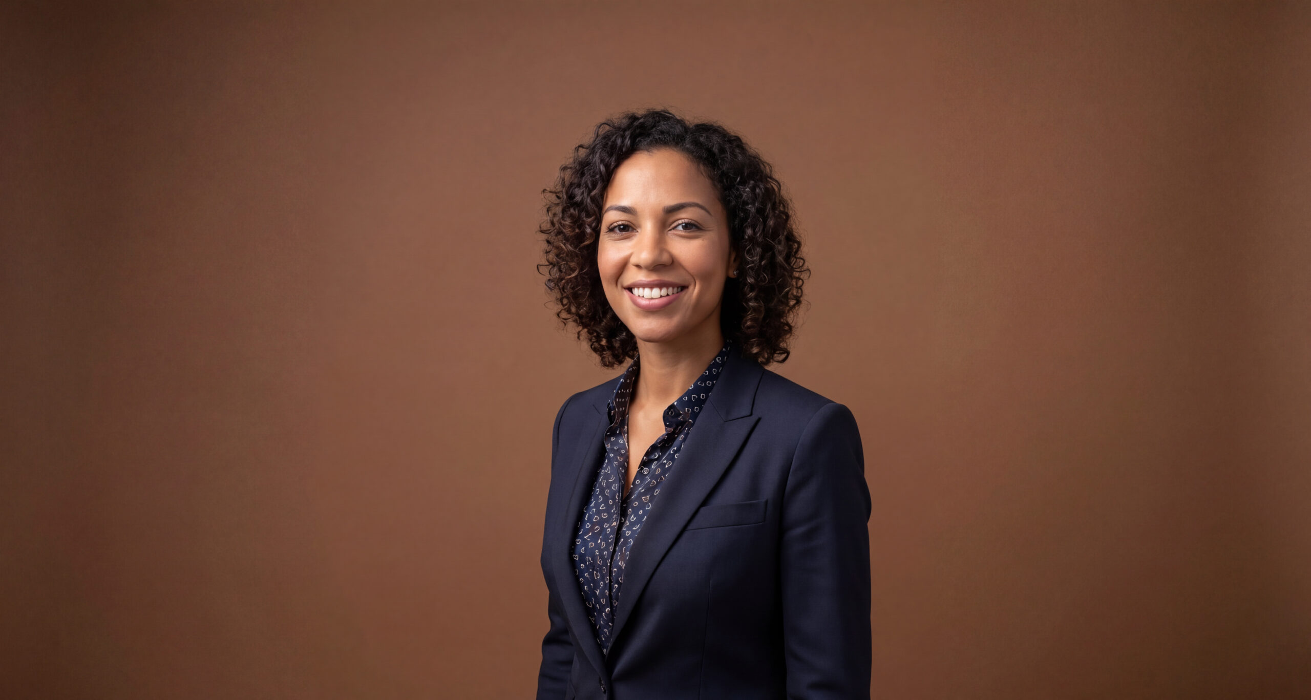 Smiling professional woman in a navy blazer stands confidently against a brown background showcasing a business attire style suitable for corporate and professional environments.