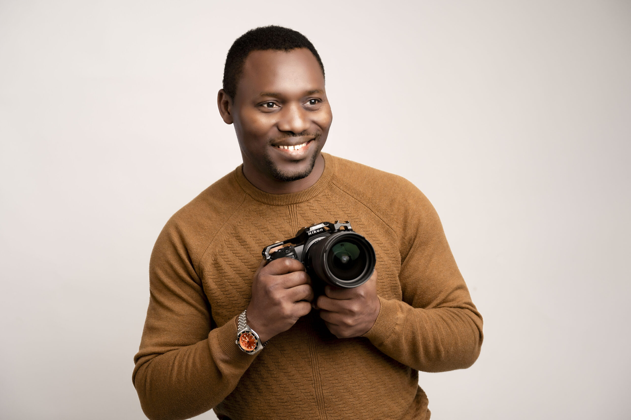 Man in a brown sweater smiling and holding a camera standing against a plain background showcasing a positive attitude and passion for photography.
