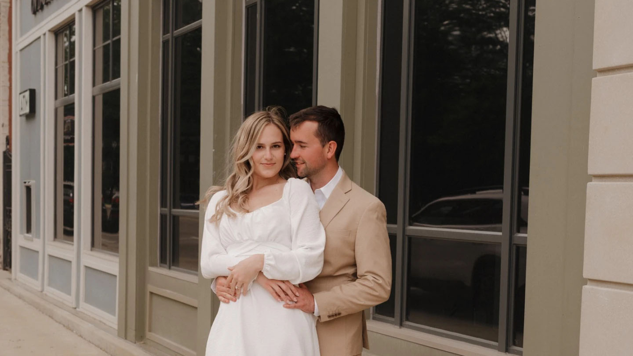 A couple embraces outside a stylish urban building on a cloudy day. The woman wears a white dress, and the man sports a beige suit, creating a sophisticated and modern look.