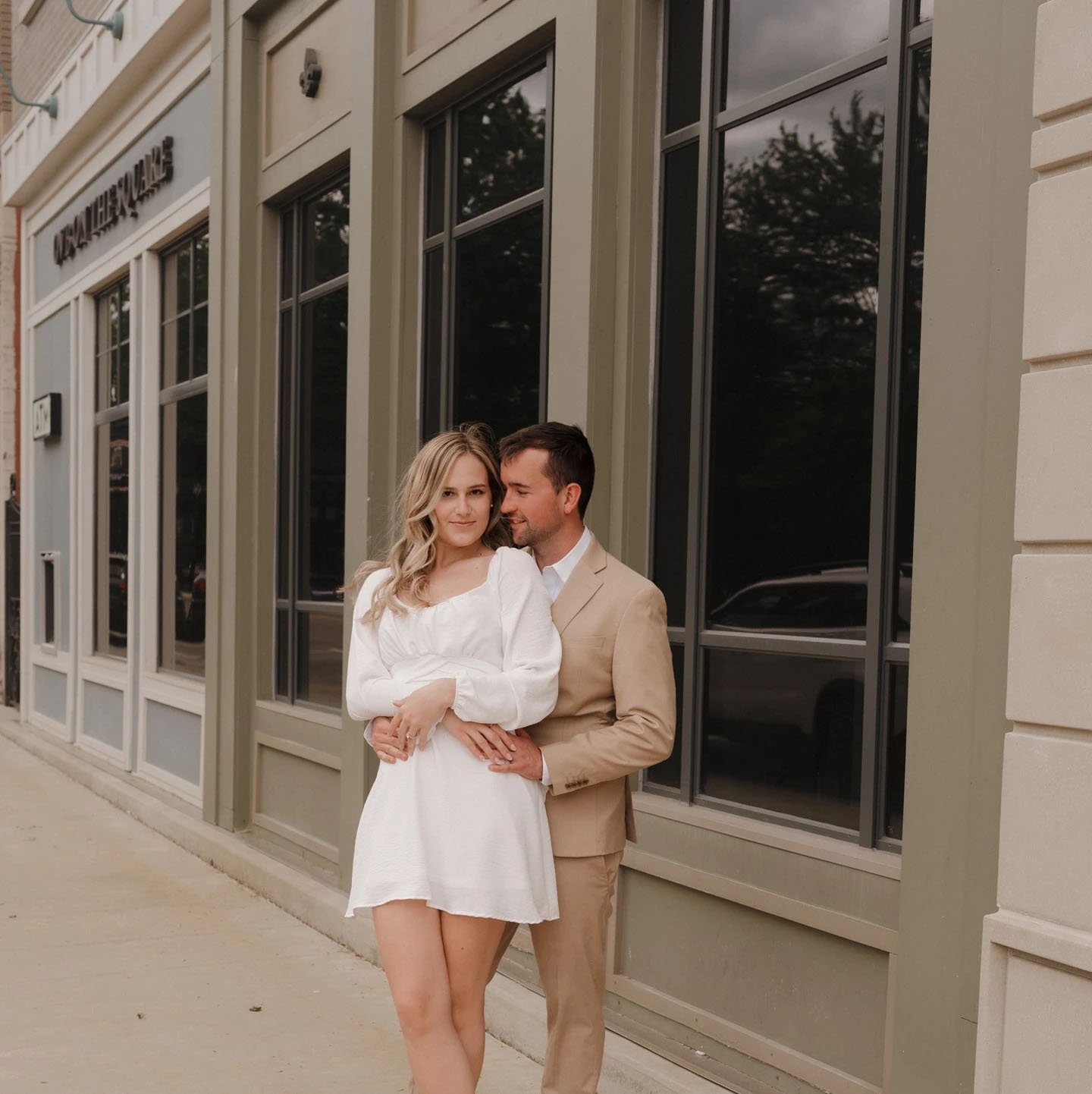 A couple embraces outside a stylish urban building on a cloudy day. The woman wears a white dress, and the man sports a beige suit, creating a sophisticated and modern look.