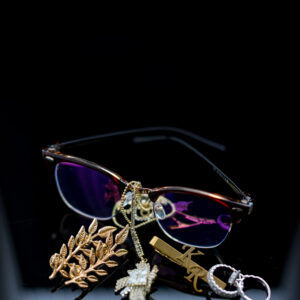 A close-up image of stylish eyeglasses with gold and silver jewelry including a brooch and pendants resting on a sleek reflective surface against a dark background.