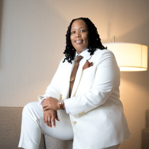 Person in a stylish white suit with a brown tie poses confidently indoors with soft lighting and modern decor creating a professional and elegant atmosphere.