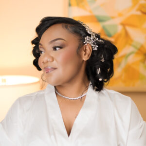Bride wearing a white robe with elegant hairstyle adorned with pearls and hair accessories in a softly lit room with abstract art in the background.