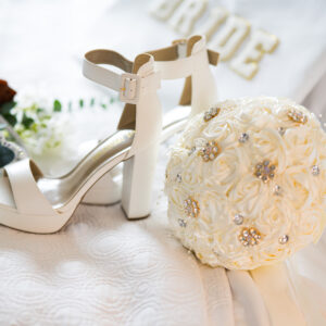 Elegant bridal accessories on display featuring white high-heeled sandals and a cream rose bouquet with sparkling accents on a white fabric backdrop for wedding elegance.