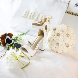Elegant bridal accessories including a white high-heeled shoe, floral decor, sunglasses, and a sign for the bride on a white bedspread, perfect for a wedding day.