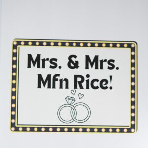 Wedding celebration sign with the text "Mrs. and Mrs. Mfn Rice" adorned with two interlocking rings under a diamond design. Perfect for announcing same-sex marriage.