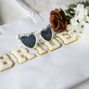 Bride tote bag with gold letters featuring stylish sunglasses and white and brown flowers on a soft bedspread perfect for wedding preparation and bridal accessories.