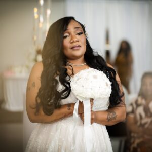 A bride in a white wedding dress holds a bouquet of white flowers. She stands elegantly in a softly lit venue with guests in the background, creating a serene atmosphere.