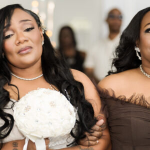 Bride holding a bouquet stands with her mother at a wedding ceremony both wearing elegant dresses and jewelry in a warmly lit venue with guests in the background.