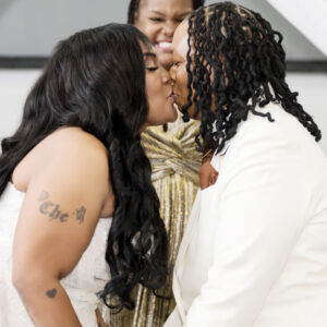 Two people share a joyful wedding kiss while holding hands dressed in elegant attire with a smiling person in the background celebrating their special union.