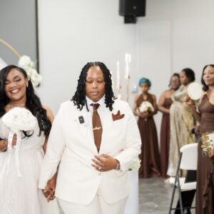 A joyful couple walks hand in hand at their wedding ceremony surrounded by bridesmaids in elegant brown dresses and white flowers in a beautifully decorated venue.