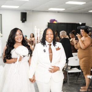 Two smiling brides walk hand in hand down the aisle in a wedding ceremony surrounded by guests capturing the joyous moment in a modern decorated venue.