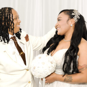 A joyful couple shares a loving gaze on their wedding day. The bride holds a bouquet, and both wear elegant attire, embodying happiness and celebration in a white-themed setting.