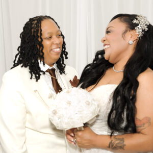 A joyful couple shares a loving moment on their wedding day. The bride holds a bouquet and wears a decorated dress while her partner smiles warmly in a white suit.