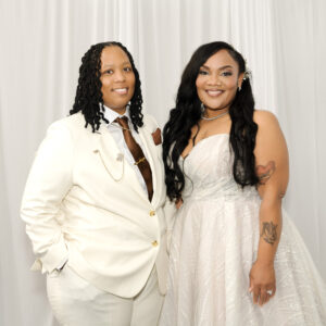 Two individuals dressed elegantly; one in a white suit and the other in a white lace gown, smiling at the camera with a white curtain backdrop, exuding happiness and style.