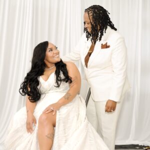 Couple in elegant white wedding attire share a joyful moment. Bride sits smiling at standing partner against a soft white backdrop capturing love and happiness on their wedding day.