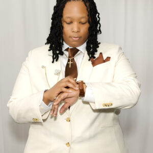 Person in a white suit with a brown tie and pocket square checking time on wristwatch showcasing formal fashion and business professionalism against a neutral background.