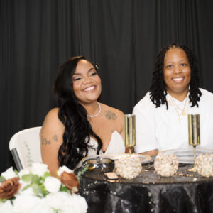 Two people smiling at an elegantly decorated table with champagne glasses and floral arrangements against a black backdrop, creating a joyful and celebratory atmosphere.
