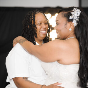 Two people embrace joyfully during a wedding ceremony with elegant attire and a beautiful backdrop. LGBTQ plus celebration highlighting love and happiness on their special day.