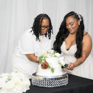 Two women smiling as they cut a wedding cake adorned with flowers. They are dressed elegantly, enjoying a special moment together at a wedding celebration.