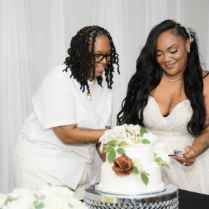 Two people joyfully cutting a wedding cake together at a beautifully decorated reception. One wears a white dress while the other wears a white shirt, both smiling warmly.