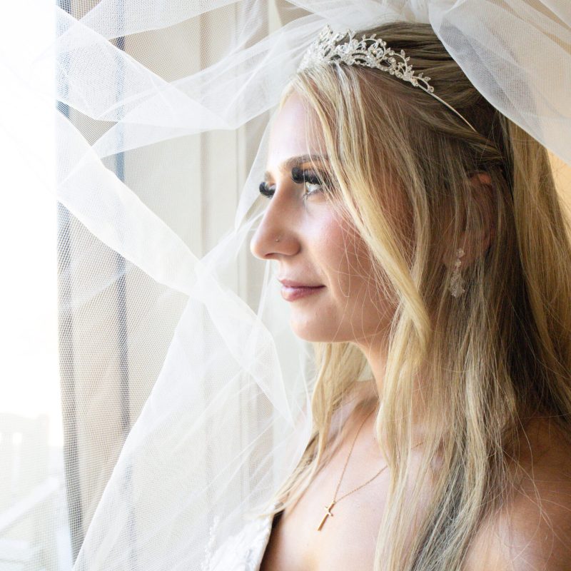 Bride gazing out of window with veil and tiara adorning her long blonde hair wearing a strapless wedding gown on her special day capturing a serene and joyful moment.