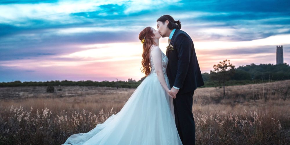 Bride and groom share a romantic kiss at sunset on a scenic outdoor landscape with dramatic skies symbolizing love and togetherness on their wedding day.